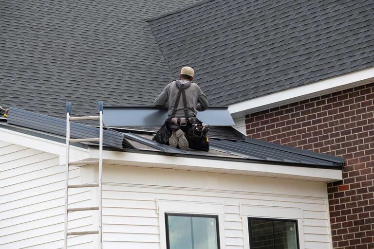 A person using a roof rake to remove snow from a house roof on a cold, clear day.